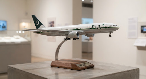 A museum-grade 1:200 scale diecast model of a Pakistan International Airlines Boeing 777-200ER aircraft in classic retro livery, mounted upon an elegant brushed curved metal arm with an American walnut wooden stand. Displayed in an archival gallery space with soft diffused studio illumination, highlighting high-precision tampo livery details and authentic landing gear assembly.