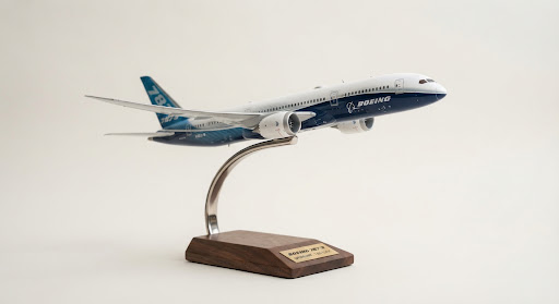 A museum-grade 1:200 scale model of a Boeing 787 Dreamliner with wings flexed, perched elegantly on a polished stainless steel curved gooseneck arm and dark walnut pedestal. Set against a clean neutral off-white museum photography background with subtle studio light reflection, emphasizing aeronautical purity and architectural grace in light mode.